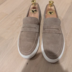 Beige mockaloafers med vit sula stl 42 - Snygga loafers i beige mocka från Goosecraft med klassisk design och vit platt sula. Skorna har rund tå och stilren ovandel utan snörning, perfekta för en clean look. Nyskick, endast använd någon gång, men något liten för mig.