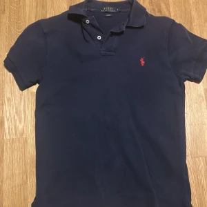Mörkblå pikétröja från Polo Ralph Lauren - Klassisk mörkblå pikétröja från Polo Ralph Lauren med röd broderad logga på bröstet. Slim fit-modell med korta ärmar och krage med två knappar. Perfekt för en stilren och sportig look.