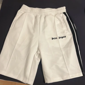 Vita shorts från Palm Angels med svarta ränder - Snygga vita shorts från Palm Angels med svarta ränder längs sidorna och logga broderad framtill. Shortsen har elastisk midja med snörning och sidofickor. Perfekta för en sportig och trendig look.