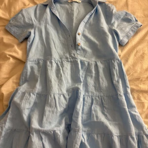 Ljusblå kort klänning från Zara - Supersöt ljusblå klänning från Zara i luftig bomull. Klänningen har korta ärmar, knappar framtill och volanger i kjolen. Perfekt för varma dagar och enkel att styla med sneakers eller sandaler.