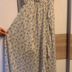 Vit långklänning med blommigt mönster och volang - Superfin långklänning i vitt med små blå blommor och guldiga detaljer. Klänningen har volang längs kjolen och ett luftigt, romantiskt fall. Perfekt för sommarens alla tillfällen!