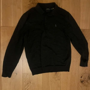 Svart långärmad pikétröja från Polo Ralph Lauren - Klassisk svart långärmad pikétröja från Polo Ralph Lauren med krage och knappar framtill. Tröjan har den ikoniska broderade loggan på bröstet och är tillverkad i ett mjukt material som passar perfekt till många tillfällen.