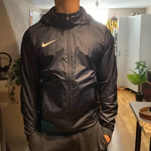 vindjacka från Nike - Säljer en mörkblå vindjacka från Nike med huva och dragkedja framtill. Jackan har lång ärm och en sportig look med vit Nike-logga på bröstet. Perfekt för utomhusaktiviteter när det blåser.