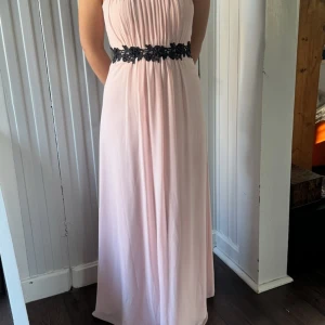 Ljusrosa axelbandslös långklänning med svart spetsdetalj - Elegant ljusrosa långklänning med axelbandslös design och svart spetsdekor i midjan. Klänningen har ett mjukt, fallande tyg och en romantisk känsla, perfekt för speciella tillfällen. Skickar snabbt!