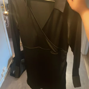 Svart omlottklänning med krage - Snygg svart omlottklänning med lång ärm och markerad krage. Klänningen har en figurnära passform och stilren design, perfekt för dig som gillar minimalistiskt mode.