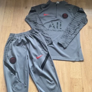 Grå Paris Saint-german tracksuit från nike - Säljer en grå långärmad Paris Saint-Germain träningströja från Nike med svarta detaljer och röd Nike-logga. PSG-emblem på bröstet och svart mönster på ärmarna. Materialet är syntetiskt och passar perfekt till träning. Den kommer också med dens vackra vackra byxor som matchar