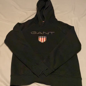 Svart hoodie från GANT - Säljer en svart hoodie från GANT med broderad logga och sköld på bröstet. Klassisk huva och mjukt material, perfekt för en avslappnad stil. Snygg detalj med röd, vit och blå rand på insidan av huvan. Pris kan diskuteras😊