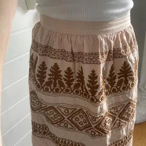 Beige/rosa kjol med brunt broderi - Säljer en beige/rosa kjol med brunt broderat mönster i form av växter och geometriska former. Kjolen har dragkedja i sidan och är lätt utställd. Perfekt för dig som gillar boho-stil och vill ha något unikt i garderoben.