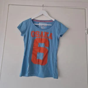 Superdry Topp - Säljer en blå t-shirt från Superdry med stort orange tryck 'OSAKA 6' på framsidan. T-shirten har rund halsringning och korta ärmar. Perfekt för en sportig och avslappnad stil.