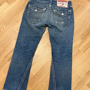 Blå jeans från True Religion med kontrastsömmar size 31-32 - Säljer ett par klassiska blå jeans från True Religion med tydliga vita kontrastsömmar och stora fickor bak med lock och knapp. Byxorna har dragkedja och raka ben. Perfekta för dig som gillar streetwear och vill sticka ut lite extra.