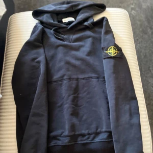Svart hoodie från Stone Island - Säljer en svart hoodie från Stone Island med klassisk logotyp-patch på ärmen. Tröjan har huva och en stor magficka framtill. Perfekt för dig som gillar streetwear och vill ha något stilrent men ändå med en tydlig detalj. Skriv pm för frågor☺️