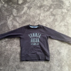 Svart sweatshirt från Gant med tryck - Fin svart sweatshirt från Gant med texten 'SUMMER BREAK BY GANT 1949' i ljusblått framtill. Tröjan har rund halsringning och långa ärmar. 