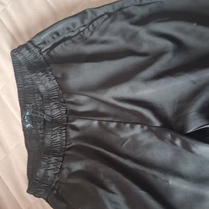 Svarta vida byxor i satin - Säljer ett par svarta, glansiga byxor från new yorke med resår i midjan. Byxorna har en loose passform och är tillverkade i ett satinliknande material som ger en lyxig känsla. Perfekta för dig som gillar bekväma och stilrena plagg.