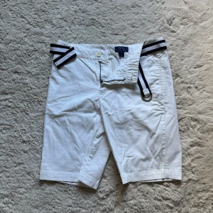 Vita shorts med bälte från Polo Ralph Lauren - Snygga vita shorts från Polo Ralph Lauren med svartvitt tygbälte. Klassisk passform med knapp och dragkedja framtill samt två bakfickor med knappar. Perfekta för sommardagar och stilrena outfits. Storleken är 18 som är 116 cm höft storlek