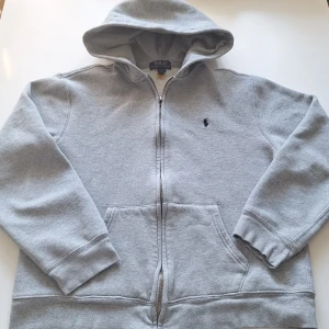 Grå hoodie från Polo Ralph Lauren - En grå hoodie från Polo Ralph Lauren med dragkedja och huva. Tröjan har broderad logga på bröstet. Riktigt snygg och passar med det mesta. Storleken är 170 men är liten i storleken. Skulle säga typ 163-170. 