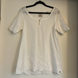 Vit blus med broderade spetsdetaljer - Säljer en vit blus från Maison Scotchmed vackra broderade spetsdetaljer längs ärmar och nederkant. Blusen har korta ärmar, rund halsringning med liten knapp och en feminin, lös passform. Perfekt för vår och sommar!