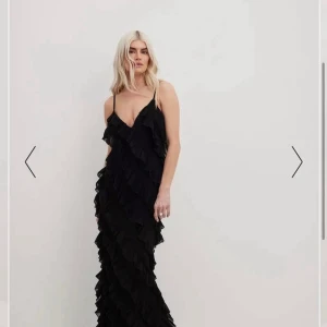 Svart volangklänning med smala axelband - Säljer en svart långklänning med smala axelband och volangdetaljer över hela klänningen. Klänningen har en v-ringad hals och en elegant, figurnära passform. Perfekt för dig som vill sticka ut med en stilren och trendig look. Använd på ett bröllop bara