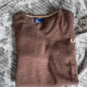 Brun tröja från Fred Perry - Säljer en brun v-ringad tröja från Fred Perry med broderad logga på bröstet. Tröjan är tillverkad i 100% ull och har långa ärmar samt ribbade muddar vid ärmslut och nederkant. Perfekt för en stilren och klassisk look.