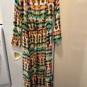 Färgglad långklänning från Zoe Baer - Säljer en långklänning från Zoe Baer med färgstarkt abstrakt mönster i grönt, gult, rosa och blått. Klänningen har lång ärm, v-ringning och knappar framtill. Perfekt för dig som gillar unika plagg med mycket färg.