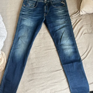 Blå jeansbyxor från Replay - Säljer ett par blå jeansbyxor från Replay med klassisk femficksdesign och snygga slitningar framtill. Byxorna har raka ben och normal passform, samt detaljerade sömmar på bakfickan. 