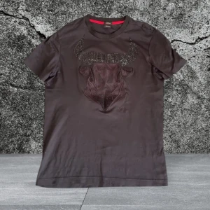 Svart t-shirt med tjurmotiv från Hugo Boss - Säljer en svart t-shirt från Hugo Boss med ett stort geometriskt tjurmotiv framtill, dekorerat med svarta pärlor. Slim fit-modell i lyxig bomull. Perfekt för dig som vill sticka ut med en unik design.