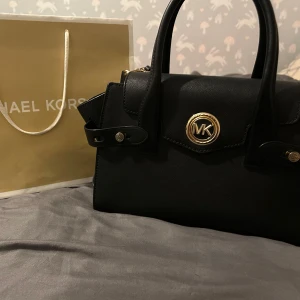 Svart handväska från Michael Kors - Säljer en stilren svart handväska från Michael Kors med guldfärgade detaljer och MK-logga framtill. Väskan har två handtag och avtagbar axelrem, perfekt för att bära på olika sätt. Klassisk design som passar till många outfits.