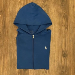 Blå hoodie från Polo Ralph Lauren - Säljer en blå hoodie från Polo Ralph Lauren med dragkedja och vit broderad logga på bröstet. Tröjan har huva och två fickor framtill. Perfekt för en avslappnad stil och enkel att matcha med jeans eller joggers.