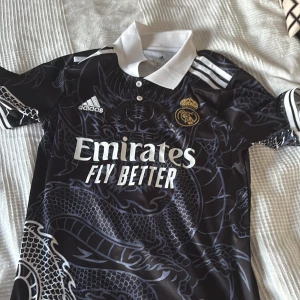 Real Madrid svart fotbollströja Adidas - Säljer en svart Real Madrid fotbollströja från Adidas med drakmönster och vita detaljer. Tröjan har vit krage, klubbmärke på bröstet och 'Emirates Fly Better' tryckt framtill. Tillverkad i AEROREADY-material för bra ventilation.