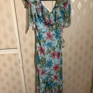 Blommig långklänning i turkos med volang - Säljer en långklänning i turkos med färgglada blommor i rosa och blått. Klänningen har volangdetaljer vid axlar och urringning samt ett luftigt, flowy material. Perfekt för sommarens tillfällen samt bröllop. Det är en liten 42a så skulle mer säga 40