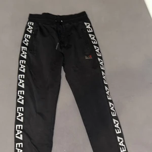 Svarta mjukisbyxor från EA7 Emporio Armani - Svarta mjukisbyxor från EA7 Emporio Armani med vita EA7-loggor längs båda benen och broderad logga på ena sidan. Byxorna har dragsko i midjan och är tillverkade i mjukt material för en bekväm passform.