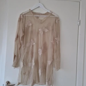 Beige transparent tunika med blommor - Säljer en beige, transparent tunika med lång ärm och v-ringning. Tunikan har stora blommiga detaljer och en luftig, flowy passform. Perfekt för lager-på-lager eller som strandplagg.