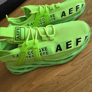 Neongröna sneakers med textdetaljer - Säljer ett par riktigt iögonfallande neongröna sneakers med svart text och unika, futuristiska sulor. Skorna har snörning och är tillverkade i syntetmaterial. Perfekta för dig som vill sticka ut med din stil!