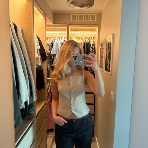 Beige korsett-topp från Zara - Snygg beige korsett-topp från Zara med smala axelband och knappar framtill. Toppen har en figurnära passform, markerad midja och en liten fejkficka på bröstet. 