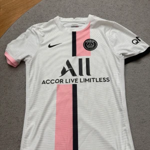 PSG vit och rosa matchtröja Nike - Paris Saint-Germain fotbollströja i vitt med en rosa och svart vertikal rand på framsidan. Broderat PSG-märke på bröstet, Nike-logga och sponsortryck. Tillverkad i lätt, ventilerande polyester. Kortärmad med rosa och svarta detaljer på ärmsluten.  SNABB AFFÄR = BÄTTRE PRIS