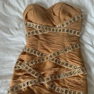Guldfärgad kort klänning med strass - Superfin kort klänning i guldfärgad med draperad design och glittriga band med stora guld- och silverfärgade stenar som korsar över hela klänningen. Axelbandslös modell med snygg sweetheart-ringning, perfekt för att sticka ut på festen