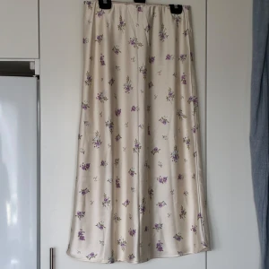 Blommig långkjol från Gina Tricot - Supersöt långkjol från Gina Tricot i ljusbeige satinliknande material med lila och gröna blommor. Kjolen sitter tajtare upptill och blir lite pösigare under knäna och är perfekt för vår och sommar. 