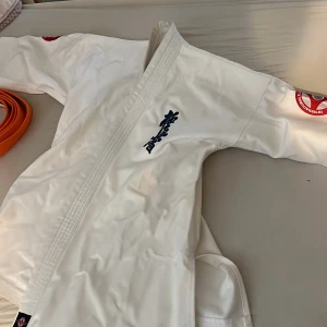 Vit karatejacka Kyokushin med brodyr - Säljer en vit karatejacka från Kanzen med Kyokushin-brodyr i svart på bröstet och röd logga på ärmen. Jackan är i kraftig bomull och har klassisk omlottknäppning. Perfekt för dig som tränar kampsport och vill ha en stilren och tålig gi. Köpt för 600kr men behöver inte längre eftersom de passar inte mig längre annars är de underbara.