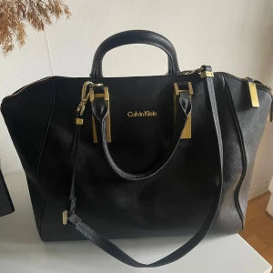 Svart skinnhandväska från Calvin Klein - Snygg svart handväska från Calvin Klein med guldfärgade metalldetaljer och dragkedja. Väskan har både handtag och axelrem, vilket gör den enkel att bära. 