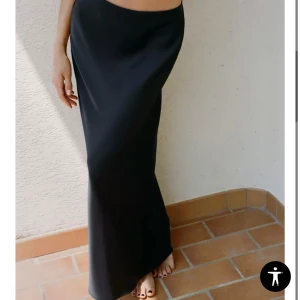 Svart satinkjol i lång modell - Supersnygg svart långkjol i satin med slät, glansig yta och rak passform. Kjolen har en minimalistisk look och faller snyggt längs kroppen. Perfekt för dig som gillar stilrena och trendiga plagg.