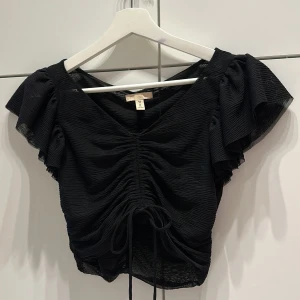 Svart croppad blus  - Svart croppad blus från H&M med v-ringning,  korta ärmar och snygg snörning framtill. Tunt, lätt transparent tyg med struktur och rynkade detaljer.  