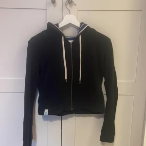 Hoodie med dragkedja och tryck - Svart croppad hoodie från WA med vit dragkedja och snörning i huvan. På ryggen står det 'NEW YORK 48' i stora vita bokstäver. Hoodien har långa ärmar och är perfekt för en sportig och avslappnad stil. Ärmlängd 56 cm, 38 cm i längden. 