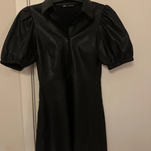 Svart kort klänning i skinnlook från Zara - Supersnygg svart kort klänning från Zara i skinnimitation med puffiga korta ärmar, krage och knappar framtill. Klänningen har en figurnära passform upptill och lätt utställd kjol. Perfekt för dig som vill sticka ut med en edgy vibe.