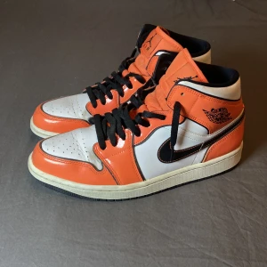 Nike Air Jordan 1 mid turf orange - Nike Air Jordan 1 sneakers i orange, vitt och svart med klassisk svart swoosh och Air Jordan-logga på sidan. Skorna har svarta snören, vit mellansula och svart bottensula. 