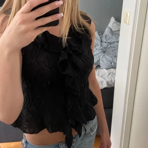 Svart ärmlös blus med volang - Supersnygg svart ärmlös blus med volangdetaljer framtill och lätt skrynklig struktur. Toppen har en croppad passform och vågig kant nertill som ger en cool och trendig look. Använd en gång💗