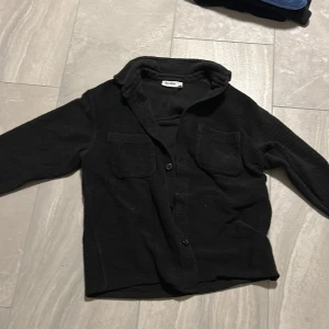 Svart overshirt från Weekday XS - Svart overshirt från Weekday i storlek XS. Skjortan har två bröstfickor, knäppning framtill och är tillverkad i ett mjukt, strukturerat tyg. Perfekt att slänga över en t-shirt för en chill look.