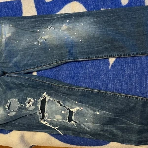 Dsquared2 blå slitna jeans herr - Riktigt snygga blå jeans från Dsquared2 med coola slitningar, stora hål och färgstänk för en edgy streetlook. Klassisk femficksmodell med orange kontrastsömmar och Dsquared2-logga vid fickan. Perfekta för dig som gillar statement-plagg.