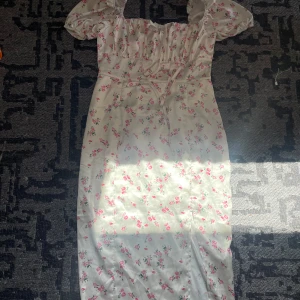 Vit blommig långklänning med puffärm - Romantisk långklänning i vit satin med rosa blommor, puffiga kortärmar och söt snörning framtill. Klänningen har en slits framtill och en smickrande passform som ger cottagecore-vibes. Perfekt för sommarens alla tillfällen!