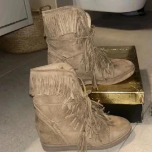 Bruna mockaboots med fransar - Snygga boots i beige mocka med coola fransdetaljer runt skaftet och längs snörningen. Skorna har kilklack och rund tå, vilket ger både höjd och komfort. Perfekta för dig som vill sticka ut med en boho-vibe och gillar detaljer som snörning och fransar.