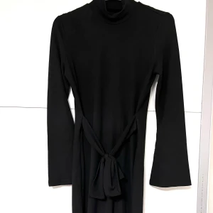 Svart långärmad klänning med knyt - En svart långärmad klänning med polokrage och knytband i midjan. Klänningen har en rak passform och är tillverkad i ett mjukt, stretchigt material.