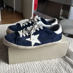 Mörkblå sneakers med vit stjärna - Snygga mörkblå sneakers från Golden Goose med vit stjärndetalj på sidan, vita snören och vit häl. Ovandelen är i mocka och skon har en klassisk platt sula. Perfekta för dig som gillar en cool och avslappnad stil.